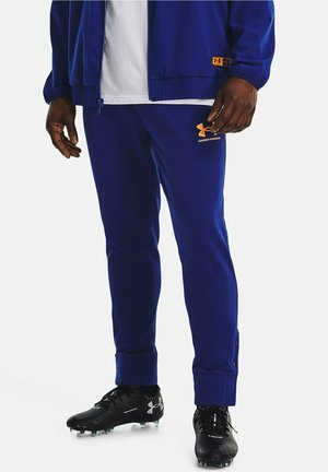 Blå træningsdragt med orange Under Armour-logo, der har en tætsiddende pasform, ribbede manchetter og trykknapper ved anklen. Sorte fodboldstøvler fuldender looket.