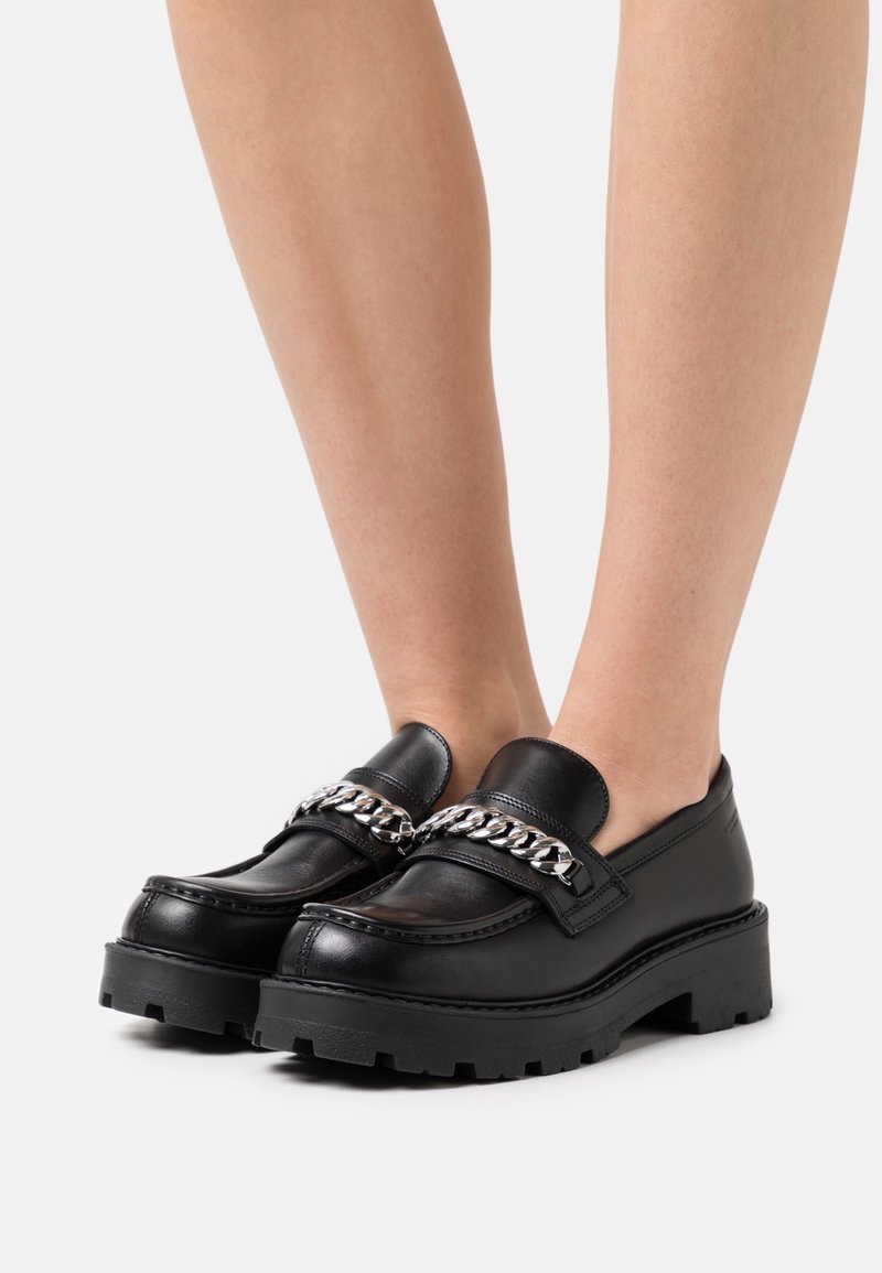 Mocassins en cuir noir avec semelle épaisse, ornés d'une chaîne argentée traversant le dessus et d'un motif de bande de roulement texturé.
