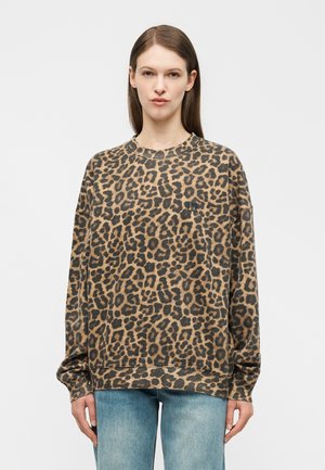 Γυναίκα με ίσια καστανά μαλλιά, που φοράει ένα καφέ και μαύρο φαρδύ φούτερ με λεοπάρ σχέδιο και ανοιχτό μπλε τζιν, στέκεται μπροστά σε λευκό φόντο.