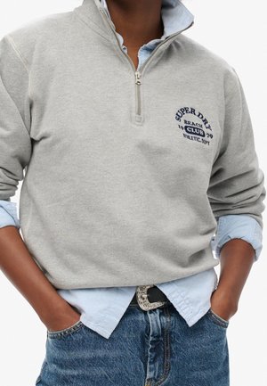 Osoba w szarym swetrze z zamkiem po ćwiartkę, nałożonym na jasnoniebieską koszulę, z rękami w kieszeniach, w niebieskich dżinsach i z ozdobną klamrą paska.