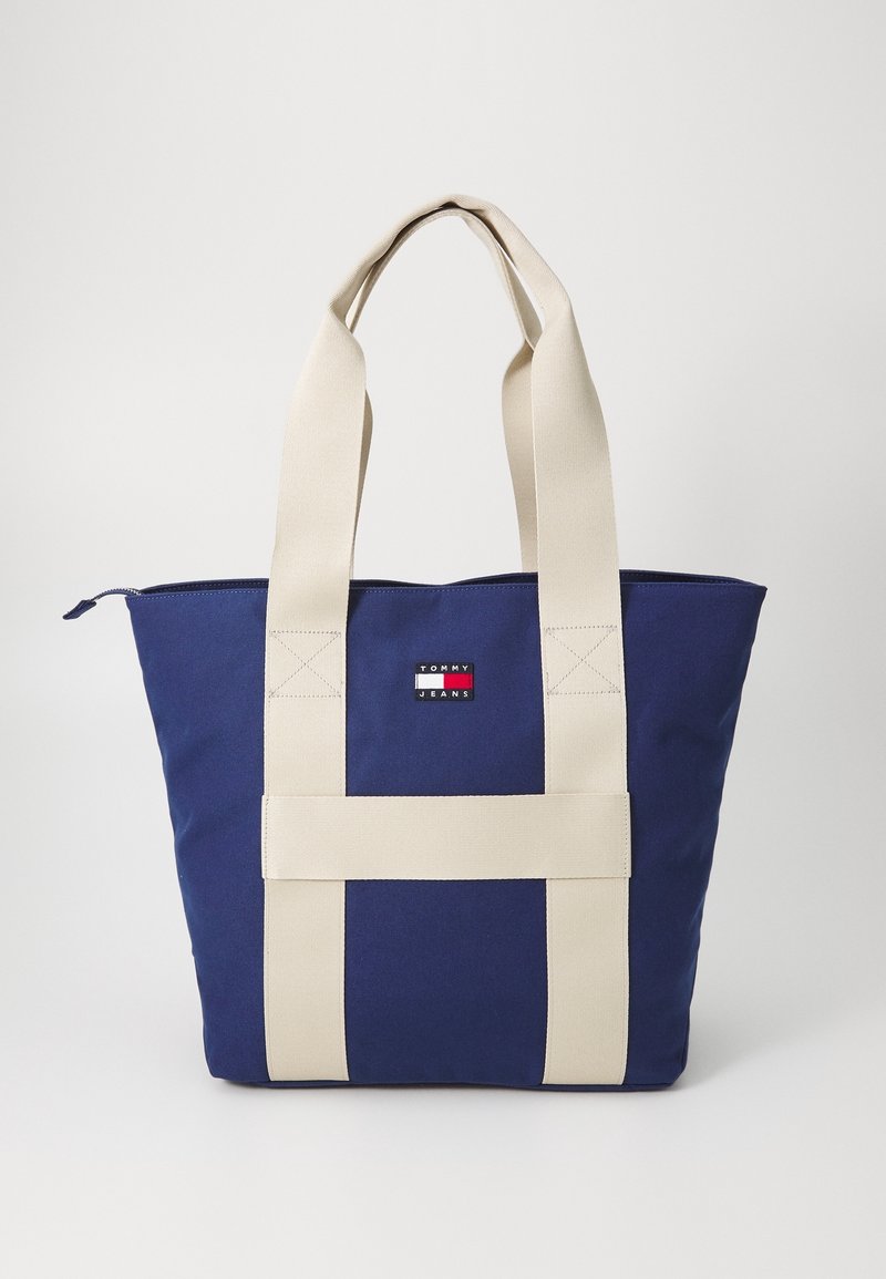 Marinblå canvas tote-väska med krämfärgade handtag, med ett logomärke på framsidan och en dragkedjestängning upptill. Robust design med förstärkta sömmar.