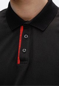 Czarna koszulka polo z ribbowanym kołnierzykiem, trzema czarnymi guzikami i czerwonym akcentem z logo wzdłuż listwy guzików. Teksturowana tkanina.