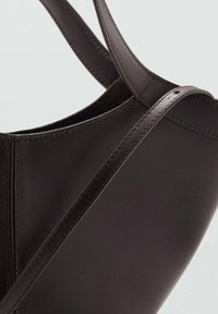 Sac à main en cuir marron foncé avec une texture lisse, un design minimaliste et des fines bretelles. La vue en biais met en évidence les détails de couture.