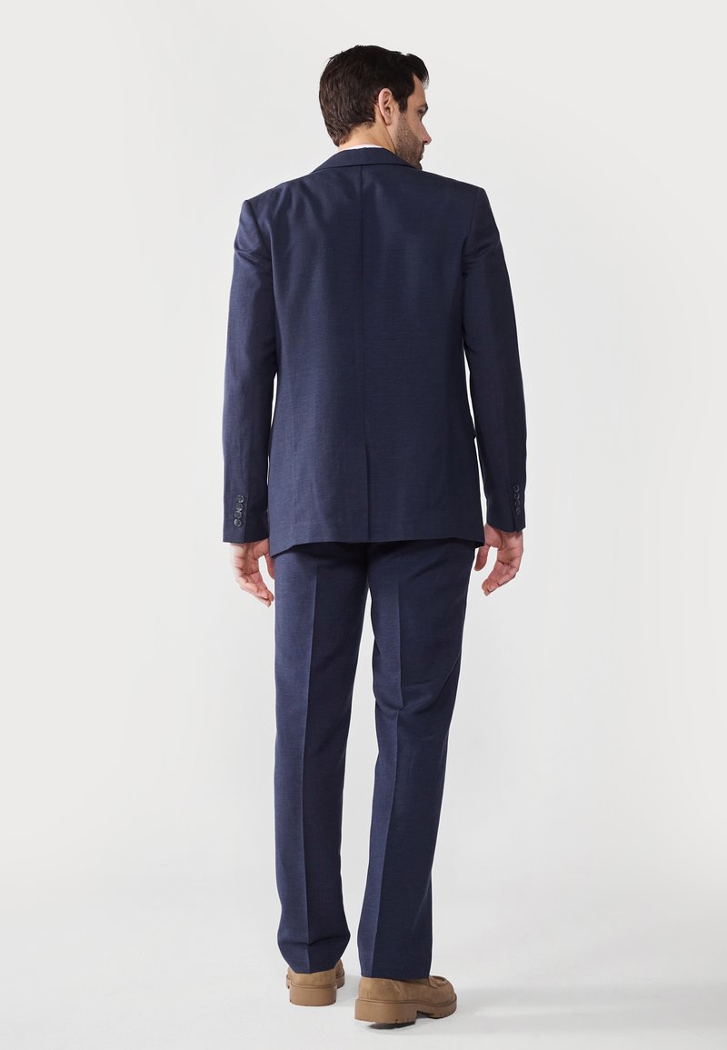 Costume bleu marine avec une veste structurée et un pantalon droit. Tissu lisse avec une texture subtile. Chaussures marron complètent la tenue.