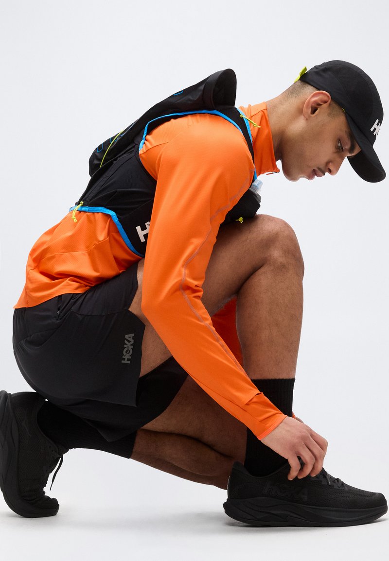 Athlet in orangefarbenem Langarmshirt und schwarzer Mütze hockt und bindet schwarzen Laufschuh, trägt schwarze Shorts und einen Trinkrucksack.