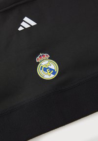 Zwarte fleece pullover met wit Adidas-logo en een blauw, geel en wit Real Madrid-embleem met een kroon bovenaan.