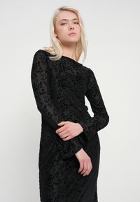 Μαύρο μακρυμάνικο φόρεμα με φλοράλ υφή, διαθέτει εφαρμοστή σιλουέτα και ελαφριά φούντα στα μανίκια. Λείο, κομψό ύφασμα.