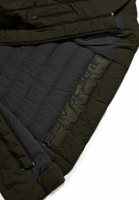 Dark green puffer jacket interior showing a quilted black lining and a glossy logo print. Features multiple zippered pockets. Texture appears soft.