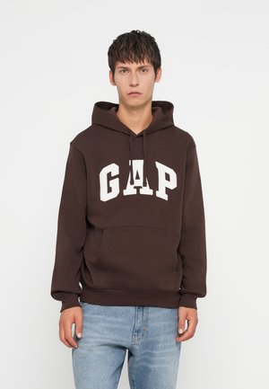 Brown hoodie made of cotton, featuring a white "GAP" logo on the front, a kangaroo pocket, and a drawstring hood. Worn with light blue jeans.