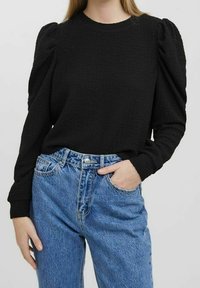 Camiseta negra de manga larga con textura y hombros abullonados, combinada con jeans de mezclilla azul claro de tiro alto con cierre de botón y cremallera.