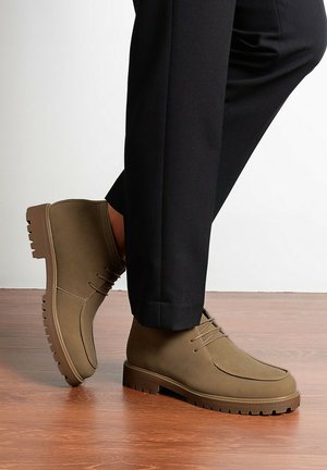 Personne portant un pantalon noir et des bottines marron à lacets, debout sur un sol en bois avec un pied levé.