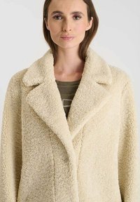 Cremefarvet, tekstureret, faux fur frakke med bred krave, enkelt knaplukning og afslappet pasform, over en mørkegrøn skjorte.