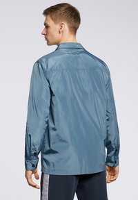 Jasnoniebieska, błyszcząca kurtka z ostro zakończonym kołnierzem, długimi rękawami i guzikami przy mankietach. Charakteryzuje się prostym krojem i minimalistycznym designem.