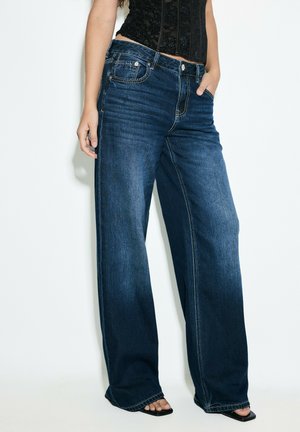 Kvinde iført mørkeblå jeans med vide ben, sort blondebluse og sorte sandaler med åben tå med den ene hånd i lommen mod hvid baggrund.