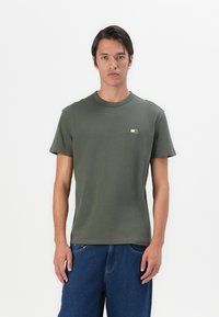 Tricou din bumbac de culoare verde măsliniu, cu mâneci scurte, de tip crew neck, și un mic design grafic pe partea stângă a pieptului. Croială obișnuită și textură moale.
