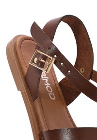 Sandalo in pelle marrone con fasce incrociate, una soletta texturizzata e un sottile dettaglio in metallo dorato. La suola è piatta con leggero plateau.