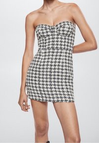 Femme portant une robe mini sans bretelles à motif pied-de-poule noir et blanc, se tenant avec un bras plié derrière son dos.