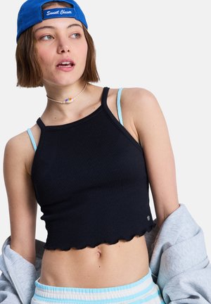 Haut court noir côtelé avec détails d'ourlet festonné, associé à des bretelles bleu clair et un sweat-shirt gris. Accessoires comprenant un collier coloré et une casquette.