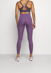 Fialová sportovní podprsenka s otevřeným zádovým designem a nastavovacími ramínky, v kombinaci s odpovídajícími legínami s vysokým pasem, doplněná o světlé růžové tenisky.