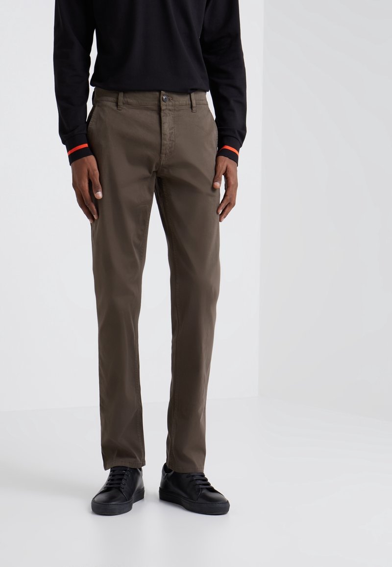 Homme portant un pantalon droit vert olive, des baskets noires et une chemise noire à manches longues avec des poignets rayés orange.
