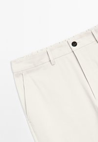 Primo piano di pantaloni beigi con chiusura a bottone, passanti per cintura e tasche laterali su uno sfondo bianco.