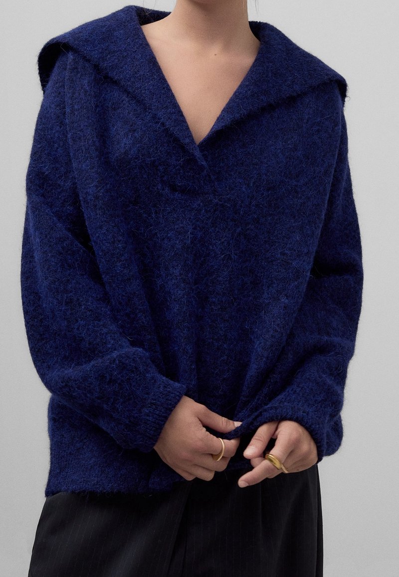 Personne portant un pull bleu foncé duveteux avec un large col, tenant l'ourlet inférieur et portant des bagues en or aux deux mains.