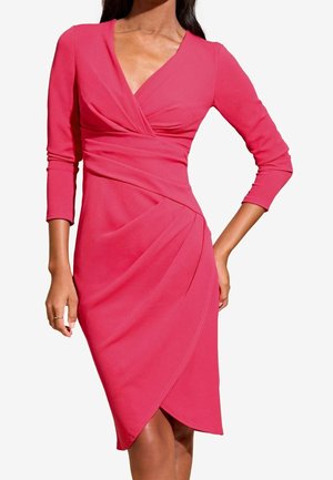 Kobieta ubrana w różową, sięgającą kolan sukienkę kopertową z rękawami do łokcia i dekoltem w kształcie litery V, stoi z jedną ręką na biodrze.