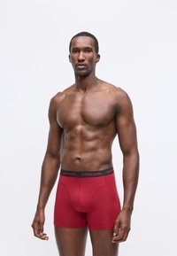Piros boxer alsónemű, puha anyagból, fekete rugalmas derékpánttal, amelyen piros szöveggel szerepel a "BJÖRN BORG" felirat. A modell semleges pózban áll.