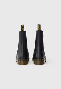 Botas de tornozelo em pele preta com superfície texturizada, costura amarela ao longo da sola e puxadores amarelos na parte de trás. Design robusto.