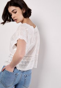 White embroidered blouse with short sleeves, featuring intricate lace patterns along the hem and neckline, worn with light blue jeans.