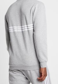 Grå sweatshirt med rund halsringning, långa ärmar och tre vita ränder över ryggen. Tillverkad av mjukt, texturerat tyg.