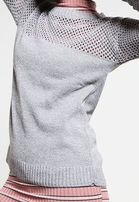 Hellgrauer Strickpullover mit strukturiertem Design, Mesh-Muster an den Ärmeln und geripptem Saum, über einem pinkgestreiften Oberteil getragen.