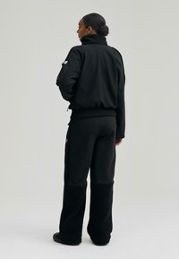 Černá bunda s vysokým límcem, hladká tkanina, doplněná černými kalhotami s jemnou, texturovanou spodní částí. Jednoduchý, funkční design.