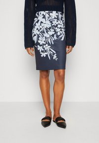 Jupe crayon florale bleu marine avec des motifs de fleurs blanches, texture lisse et design ajusté. Associée à des ballerines noires à bout pointu.