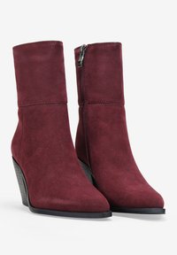 Botas de tobillo de ante burdeos con punta afilada, un diseño estilizado y cremallera lateral. Presentan un tacón negro apilado para mayor altura.