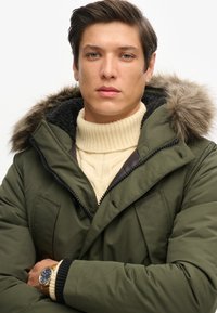 Parka verde oliva con capucha con borde de piel, cierre de cremallera y bolsillos frontales. Llevada sobre un suéter de punto color crema, con textura acanalada.