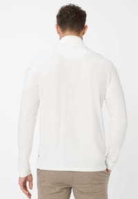 Uomo visto da dietro che indossa una camicia bianca a maniche lunghe e pantaloni beige chiaro con texture, in piedi contro uno sfondo bianco uniforme.