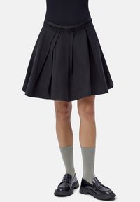 Woman wearing a pleated black skirt, gray ribbed mid-calf socks, and black leather loafers, standing against a white background.
