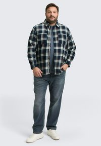 Flanellen geruite shirt in marineblauw, wit en rode tinten; heeft twee borstzakken en knopen aan de voorkant, gedragen over een marineblauw T-shirt en blauwe spijkerbroek.
