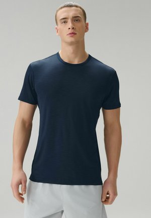 Junger Mann mit kurzen Haaren, der ein dunkelblaues T-Shirt mit Rundhalsausschnitt und kurzen Ärmeln sowie eine hellgraue kurze Hose trägt, vor einem einfarbigen Hintergrund steht.