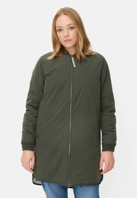 Olijfgroene lange jas met een rits aan de voorkant, zijzakken en ribgebreide manchetten. Gladde textuur, lichte stof en een losse pasvorm.