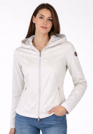 Donna con giacca grigia chiara con cappuccio e zip, con toppa logo sulla manica sinistra e jeans blu, che posa con una mano in tasca.