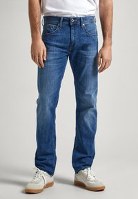 Vaqueros de mezclilla azules con un corte recto, que presentan un diseño clásico de cinco bolsillos, costuras visibles y una cintura de altura media. Combinados con zapatillas blancas.