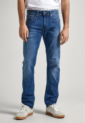 Vaqueros de mezclilla azules con un corte recto, que presentan un diseño clásico de cinco bolsillos, costuras visibles y una cintura de altura media. Combinados con zapatillas blancas.
