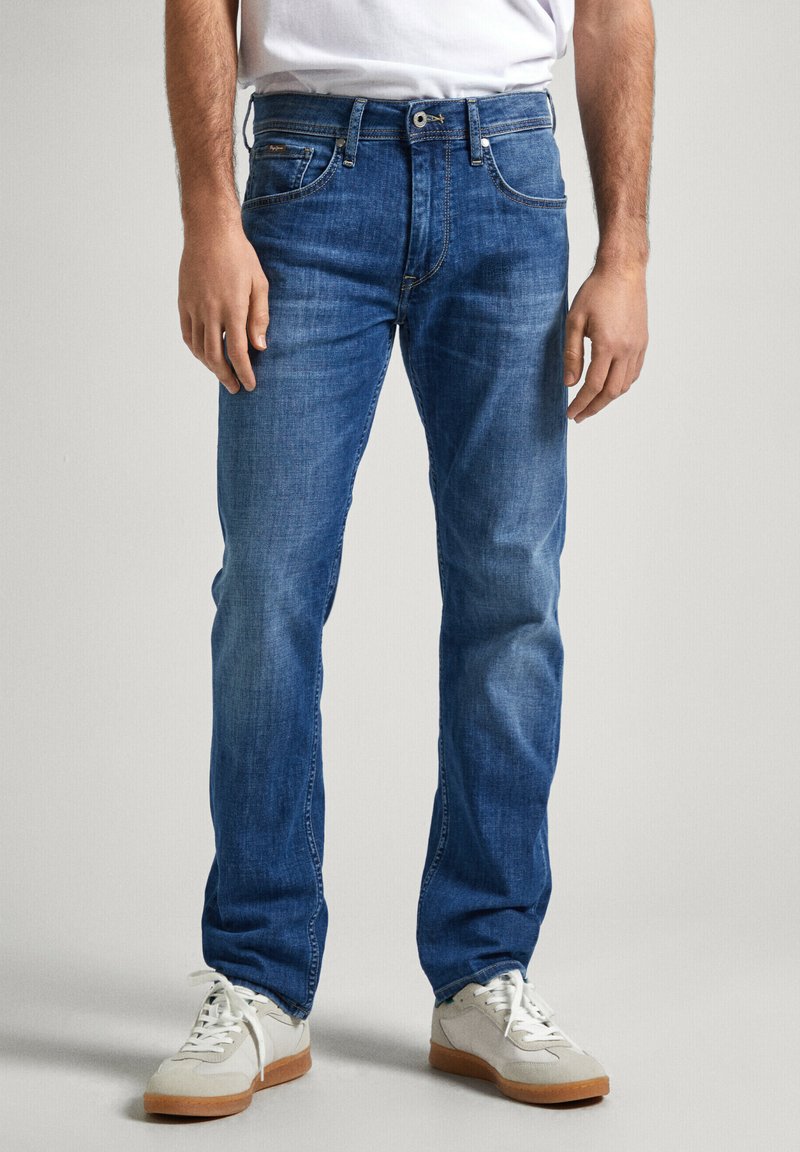 Vaqueros de mezclilla azules con un corte recto, que presentan un diseño clásico de cinco bolsillos, costuras visibles y una cintura de altura media. Combinados con zapatillas blancas.