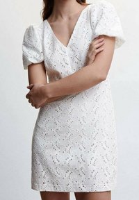 Robe courte blanche avec manches bouffantes, col en V et motifs en dentelle découpée complexes. Le tissu semble texturé et léger.