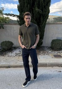 Mladý muž s krátkymi vlasmi kráča vonku, má na sebe zelené polo tričko, tmavé džínsy, čierne tenisky, ruky v džepoch a v pozadí sú stromy.