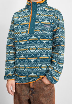 La personne porte un pull polaire bleu et orange à motifs avec des boutons-pression et une poche poitrine zippée, associé à un pantalon marron.