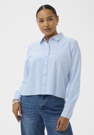 Femme aux cheveux plaqués en arrière, portant une chemise bleue claire boutonnée et un jean bleu, debout devant un fond uni.