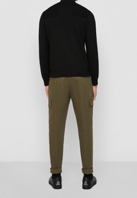 Black knit turtleneck sweater with a textured yoke, paired with olive cargo pants featuring side pockets and rolled cuffs, worn with black shoes.